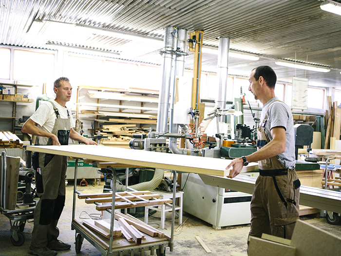 Branche Handwerk; Tischlerwerkstatt mit zwei Tischlern die gerade eine Platte auf einen Rollwagen heben;